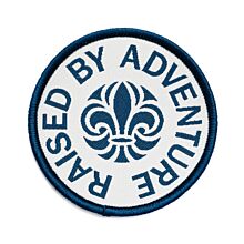 Runt märke med blå langetterad kant och vit bakgrund med scoutsymbolen på och runt den texten Raised By Adventure