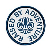 Runt märke med blå langetterad kant och vit bakgrund med scoutsymbolen på och runt den texten Raised By Adventure