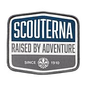 Märke med grå langetterad kant, vit bakgrund och blå detalj med texten Scouterna Raised By Adventure since 1910 och scoutsymbolen på